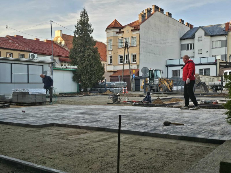Widok na plac miejski w trakcie prac brukarskich. Duża część terenu została już wyłożona szarą kostką brukową. Na pierwszym planie, na fragmencie nieutwardzonego jeszcze piasku, leży młotek brukarski. Po lewej stronie robotnik pochyla się nad paletą z kostką, a po prawej stronie kadru idzie mężczyzna w jasnych butach i czerwonej kurtce. W tle widoczne są wielopiętrowe kamienice o zróżnicowanej architekturze