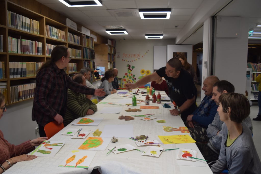 Perspektywiczne ujęcie długiego, białego stołu w bibliotece, wokół którego kilkanaście osób uczestniczy w warsztatach plastycznych. Na stole leżą gotowe rysunki o tematyce jesiennej, pędzle, liście oraz palety z farbami. Prowadzący w koszuli w kratę rozmawia z uczestnikami po lewej stronie, podczas gdy inny mężczyzna na środku sali pochyla się nad stołem, podając komuś pędzel. W tle widać dekorację z napisem „JESIEŃ”