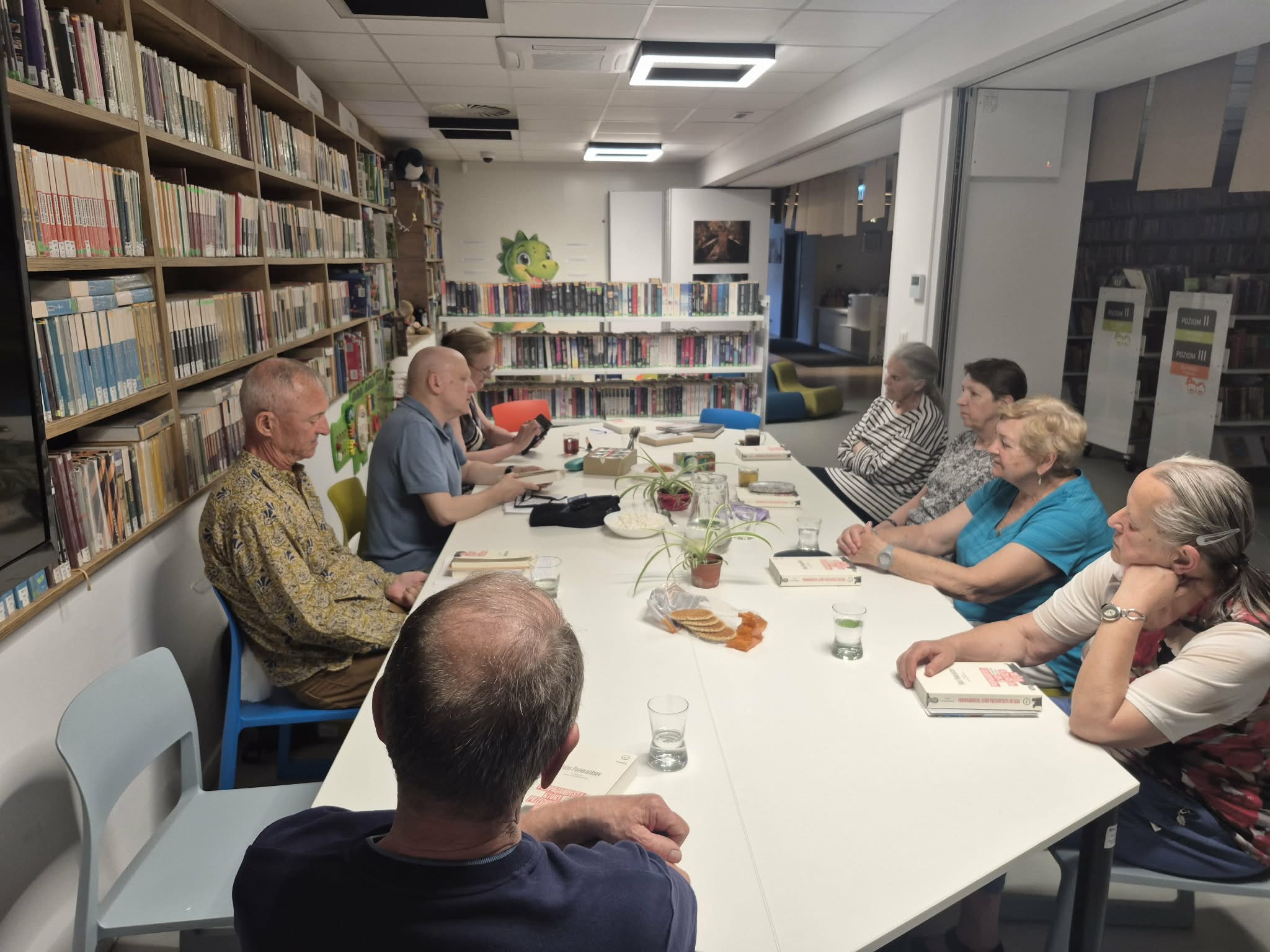 Grupa siedmiu osób – kobiet i mężczyzn – siedzi wokół białego stołu w sali bibliotecznej podczas dyskusji o książkach. Na stole leżą otwarte egzemplarze publikacji, stoją szklanki z wodą oraz talerzyki z drobnymi przekąskami. Po lewej stronie widoczne są wysokie, drewniane regały wypełnione książkami