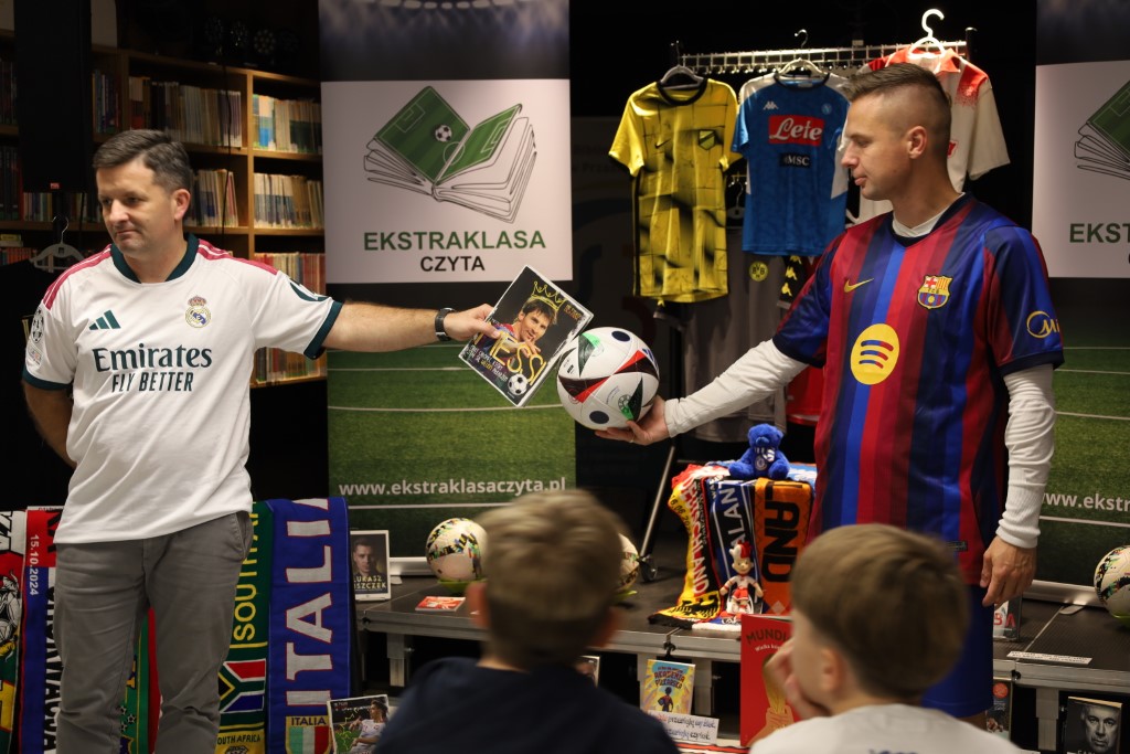 Dwaj mężczyźni w sportowych koszulkach prezentują przed zgromadzonymi dziećmi akcesoria piłkarskie. Jeden wyciąga rękę z książką o Messim, a drugi trzyma na dłoni piłkę do gry. W tle widać banery projektu „Ekstraklasa czyta” oraz wieszaki z koszulkami i szalikami klubowymi