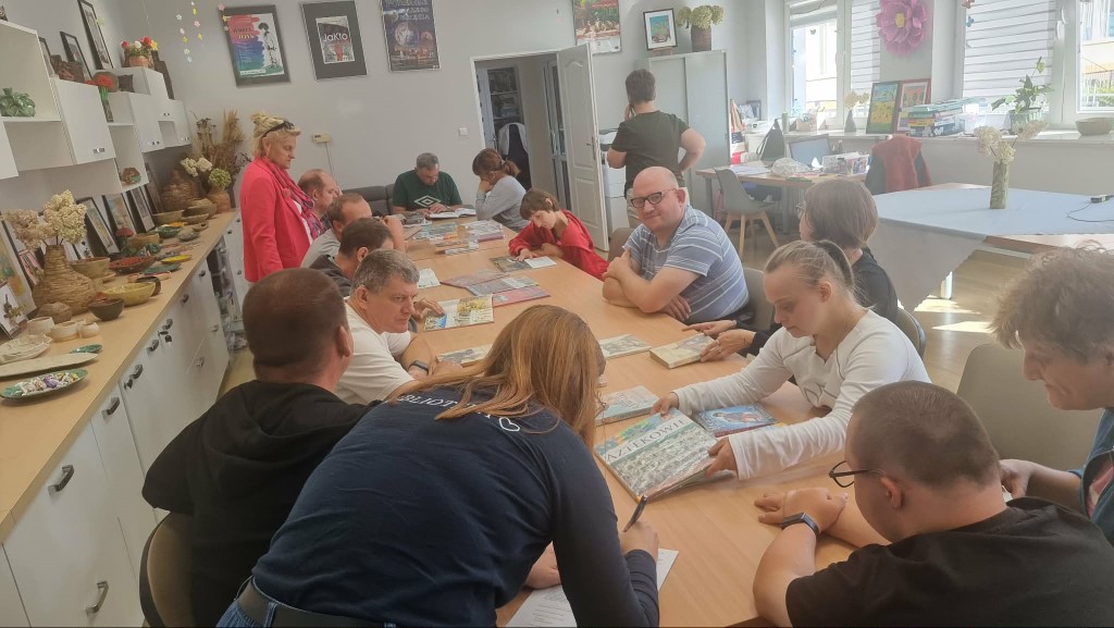 Szerokie ujęcie z góry na grupę kilkunastu osób siedzących przy długim, drewnianym stole w jasnej świetlicy. Uczestnicy, w tym osoby dorosłe i młodzież, przeglądają rozłożone na stole książki i ilustracje. Na pierwszym planie widać kobietę w ciemnym ubraniu. W tle widać białe szafki z wystawą rękodzieła (ceramika, suche bukiety) oraz plakaty na ścianach. Panuje swobodna atmosfera skupienia