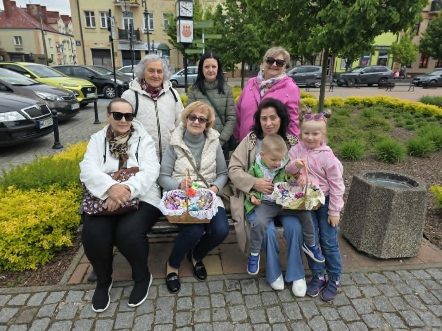 Grupa sześciu uśmiechniętych kobiet i dwoje dzieci pozuje do zdjęcia na rynku w Przasnyszu. Część osób siedzi na ławce, pozostałe stoją za nimi. Kobiety trzymają wiklinowe koszyki z drobnymi upominkami. W tle widoczne są zaparkowane samochody, kamienice oraz miejska zieleń