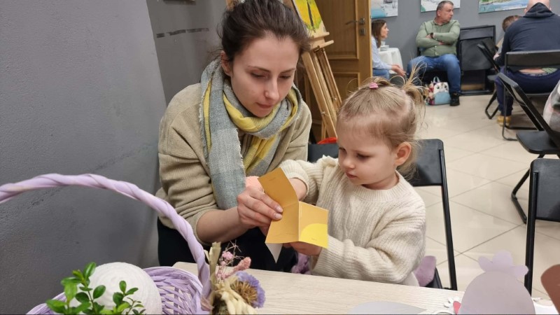Na pierwszym planie widać kobietę, która pomaga małej dziewczynce robić wielkanocną pracę plastyczną. Obie siedzą przy stoliku, naprzeciw nich stoi wiklinowy kosz z ozdobą. Kobieta najprawdopodobniej jest mamą dziewczynki