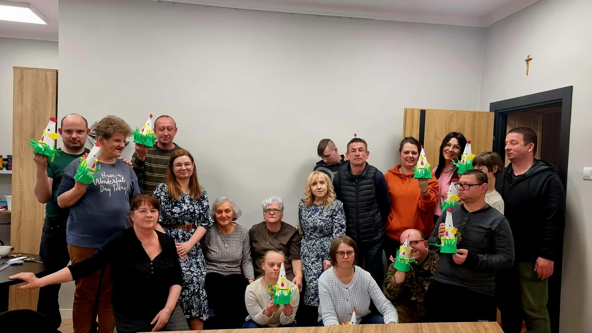 Grupa uczestników Środowiskowego Domu Samopomocy pozuje z bibliotekarkami do zdjęcia. Część osób na fotografii trzyma w rękach wykonane podczas warsztatów barwne koguty – tradycyjne ozdoby wykonane z papieru i bibuły