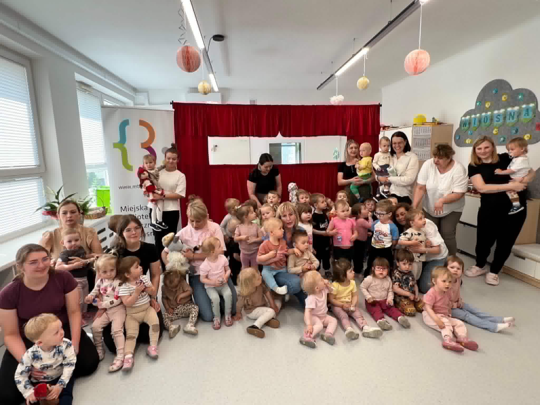 Pamiątkowe, grupowe zdjęcie w jasnym pomieszczeniu bibliotecznym lub przedszkolnym. Kilkadziesięcioro małych dzieci (w wieku żłobkowym i przedszkolnym) siedzi lub stoi w towarzystwie kilku dorosłych kobiet. W tle widoczna jest czerwona, materiałowa kurtyna, lampiony pod sufitem oraz baner z logo Miejskiej Biblioteki Publicznej