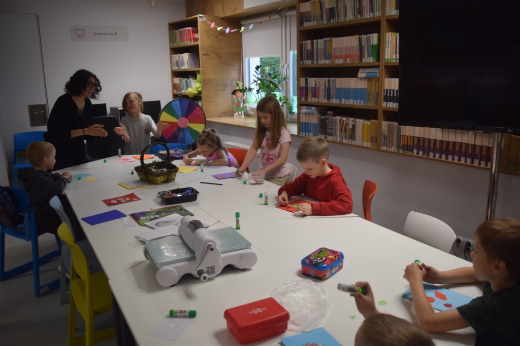 Grupa dzieci siedzi przy dużym białym stole w bibliotece i wykonuje kartki okolicznościowe metodą scrapbookingu. Na stole leżą kolorowe papiery, kleje w sztyfcie i maszynka do wycinania wzorów. W tle widać regały z książkami oraz uśmiechniętą instruktorkę rozmawiającą z dziećmi