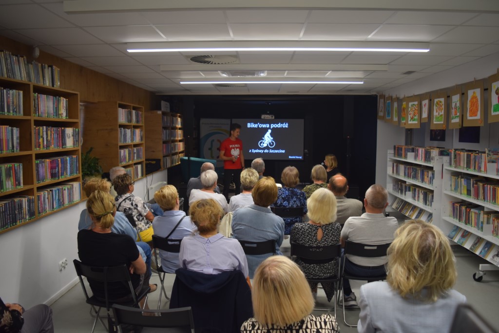Szerokie ujęcie z głębi sali bibliotecznej na spotkanie autorskie z pisarzem Przemysławem Piotrowskim. Pisarz i moderatorka siedzą na scenie na niebieskich fotelach, a przed nimi zgromadzona publiczność siedząca w rzędach krzeseł. Regały z książkami otaczają salę z obu stron, a na ścianach wiszą rysunki. Na ekranie w tle widoczny slajd tytułowy