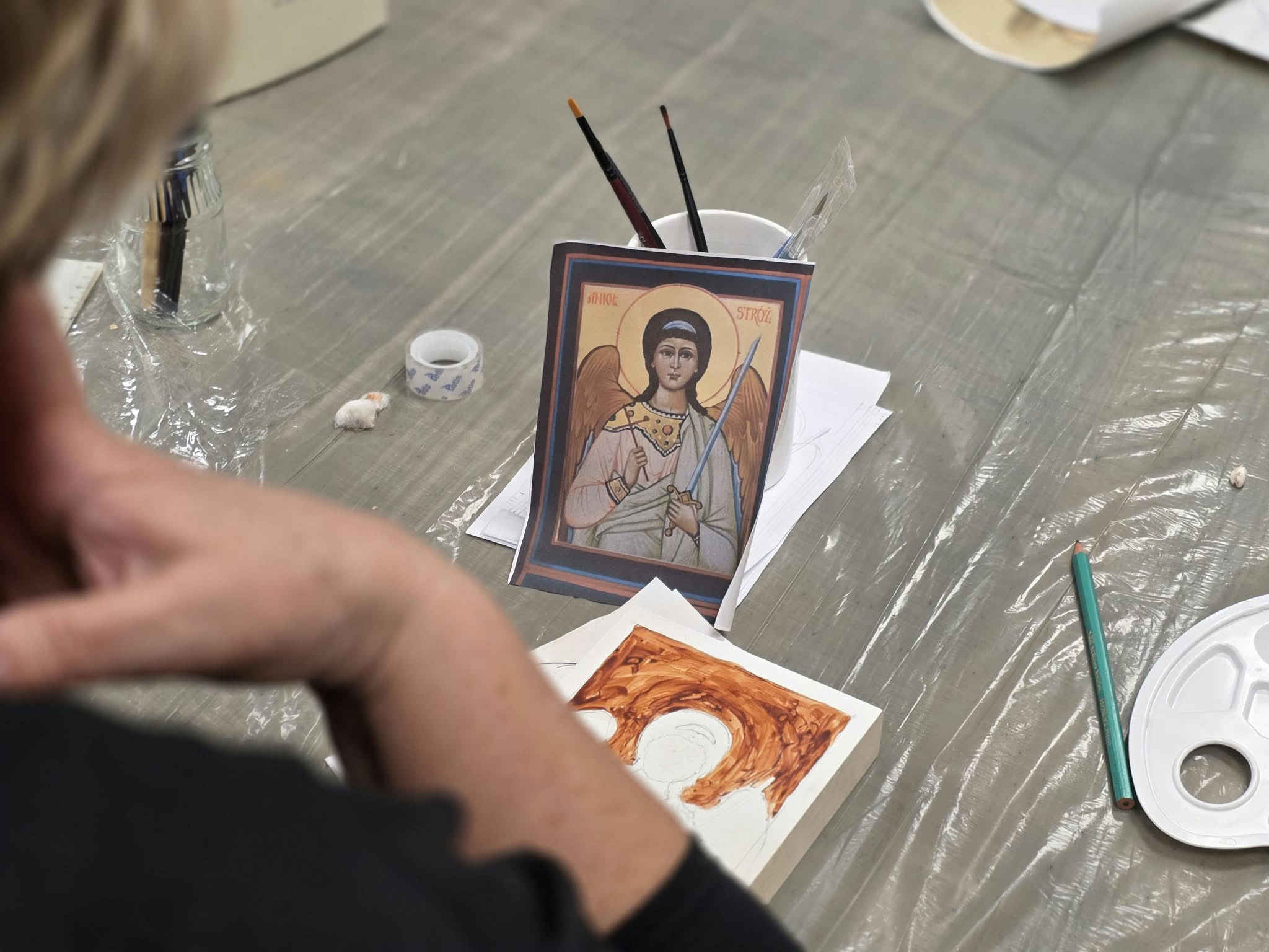 Spojrzenie zza pleców uczestniczki warsztatów na stół, na którym leży wzór ikony z Aniołem Stróżem w kubku oraz podkładka z rozpoczętym malunkiem