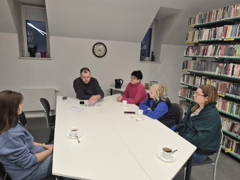 Pięć osób uczestniczy w spotkaniu przy dużym, jasnym stole w pomieszczeniu bibliotecznym ze skosami. Mężczyzna w ciemnej bluzie siedzi w centrum, opierając się o stół i rozmawiając z pozostałymi uczestniczkami. Po lewej stronie kadru widoczny jest wysoki regał z książkami, a na białej ścianie w tle wisi okrągły zegar wskazujący godzinę za dziesięć piąta