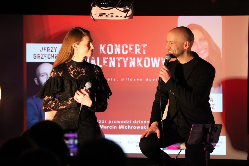 Natalia Srokocz i Jerzy Grzechnik na scenie podczas występu duetu. Obydwie osoby trzymają mikrofony i patrzą na siebie z uśmiechem. W tle wyświetlany jest czerwony slajd z napisem „Koncert Walentynkowy – duety, miłosne duety...”