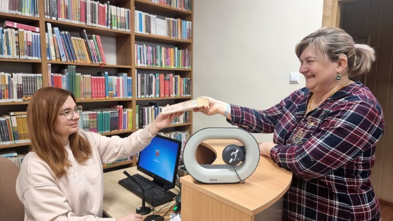 Pracownica biblioteki podaje książkę czytelniczce przy ladzie, na której stoi pętla indukcyjna dla osób słabosłyszących