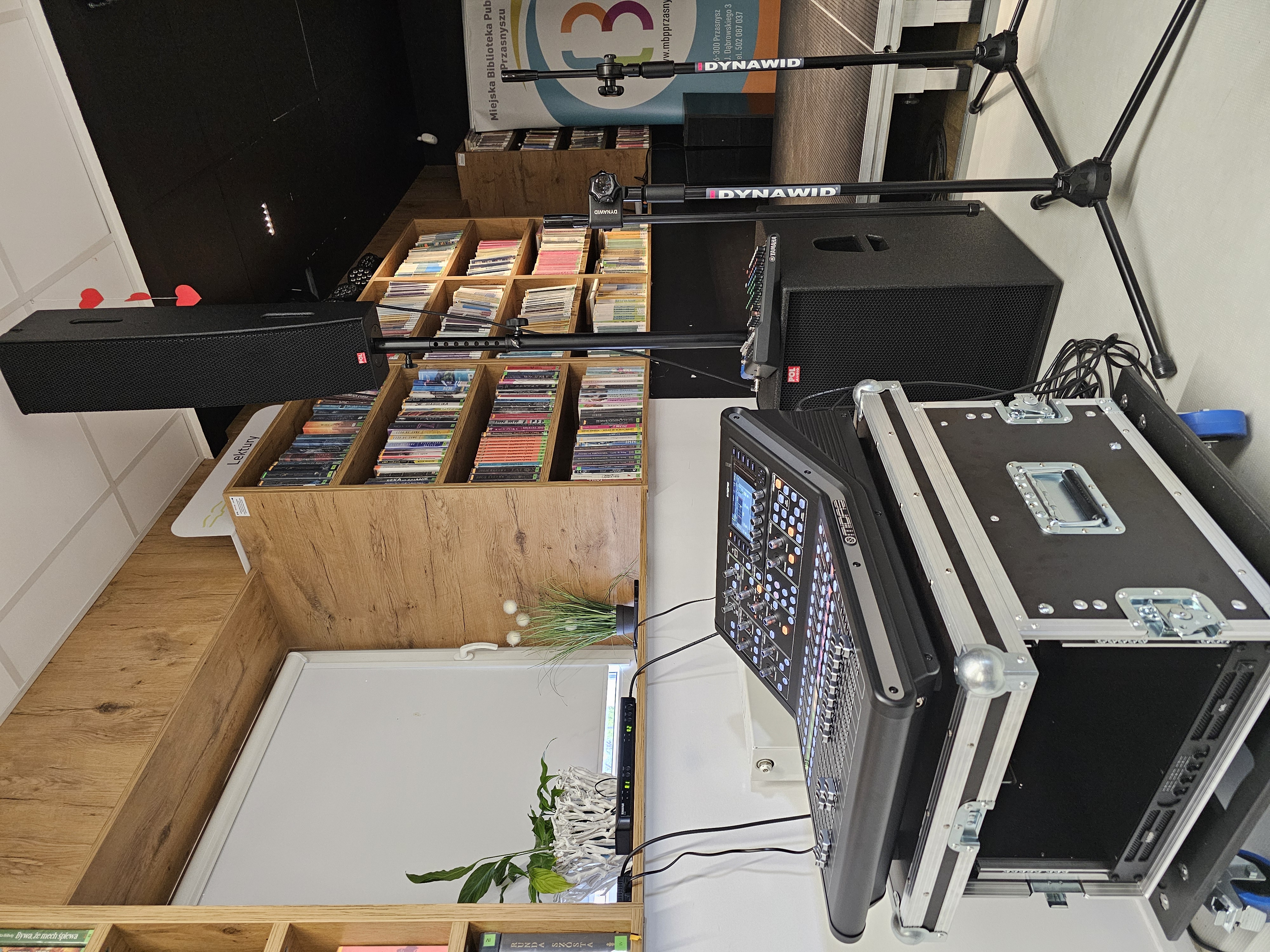 Profesjonalny sprzęt nagłośnieniowy ustawiony w pomieszczeniu bibliotecznym. Na pierwszym planie widoczny jest duży, czarny cyfrowy mikser dźwięku marki Midas, umieszczony na otwartej skrzyni transportowej typu flight case z kółkami. Po prawej stronie stoi wysoka, czarna kolumna głośnikowa na statywie, umieszczona nad dużym subwooferem. Obok znajdują się dwa statywy mikrofonowe marki Dynawid. W tle widoczne są jasne drewniane regały z lekturami i książkami, fragment sceny oraz baner Miejskiej Biblioteki Publicznej w Przasnyszu