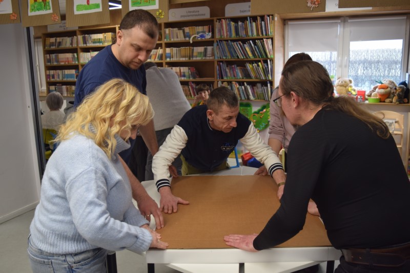 Grupa pięciu osób współpracuje przy białym stole w jasnej sali bibliotecznej. Wspólnie dociskają i wyrównują duży arkusz brązowej tektury. W tle widoczne są regały z książkami oraz zawieszone pod sufitem dziecięce rysunki o tematyce jesiennej