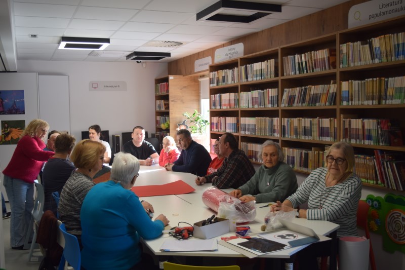 Grupa kilkunastu osób siedzi wokół połączonych białych stołów w jasnej sali bibliotecznej z regałami pełnymi książek. Na stole leżą materiały plastyczne: czerwony karton, klej na gorąco, nożyczki oraz sznurki. Uczestnicy rozmawiają ze sobą, przygotowując się do pracy manualnej