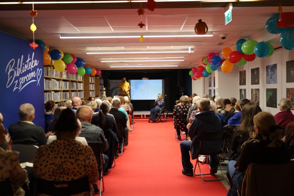 Widok od tyłu na widownię zgromadzoną w bibliotece na spotkaniu z podróżnikiem Kamilem Mąkoszą. Pośrodku sali biegnie czerwony dywan, przy regałach i na suficie wiszą kolorowe balony, a na scenie z tyłu widać prelegenta stojącego obok ekranu oraz dyrektor biblioteki