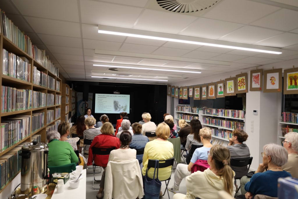 Perspektywa z boku sali bibliotecznej, ukazująca liczną grupę dorosłych osób słuchających wykładu. Nad regałami po prawej stronie wiszą oprawione rysunki dziecięce. Na pierwszym planie po lewej stronie stoi stolik z cateringiem, na którym umieszczono duży, metalowy termos gastronomiczny na napoje oraz białe filiżanki