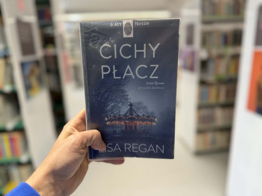 Dłoń trzyma książkę autorstwa Lisy Regan pod tytułem „Cichy płacz”. Okładka jest utrzymana w mrocznej, granatowej tonacji i przedstawia oświetloną karuzelę w mglistym parku nocą. Na górze widnieje napis „Ślady zbrodni” oraz symbol odcisku palca, a pod tytułem dopisek „Josie Quinn prowadzi śledztwo”. W tle widać rozmazane regały biblioteczne
