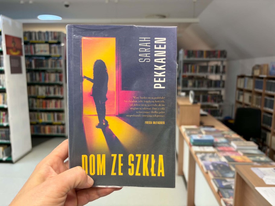 Książka „Dom ze szkła” autorstwa Sarah Pekkanen trzymana w dłoni na tle wnętrza biblioteki. Na okładce widoczna jest sylwetka dziewczynki stojącej w jasnych, otwartych drzwiach. W tle znajdują się regały z książkami oraz stoliki z wyeksponowanymi publikacjami.