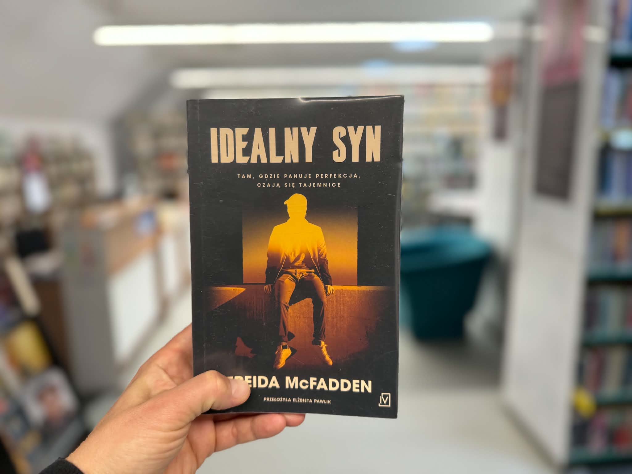 Męska dłoń trzyma książkę Freidy McFadden pt. Idealny syn z podtytułem Tam, gdzie panuje perfekcja, czają się tajemnice. Okładka przedstawia mroczną sylwetkę mężczyzny w czapce z daszkiem, siedzącego tyłem do jasnego, pomarańczowego źródła światła. Tło zdjęcia jest rozmyte i przedstawia regały w bibliotece