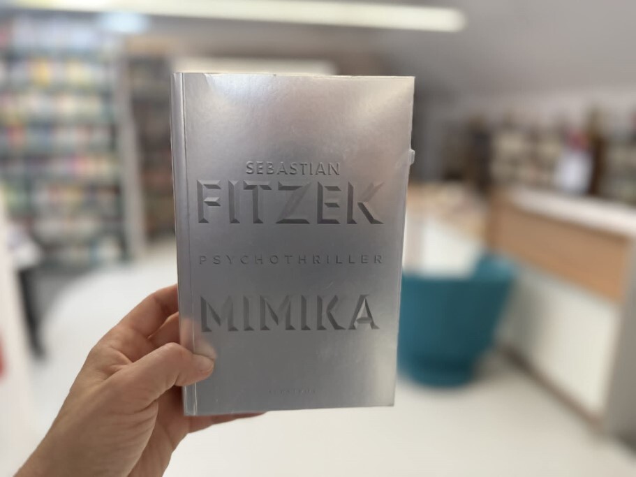 Dłoń trzymająca srebrną książkę autorstwa Sebastiana Fitzka pt. „Mimika”. Na okładce widnieje napis „Sebastian Fitzek”, poniżej znajduje się słowo „Psychothriller”, a na dole duży napis „Mimika”. Całość jest utrzymana w metalicznej, srebrnej kolorystyce. Tło zdjęcia jest rozmytym wnętrzem biblioteki