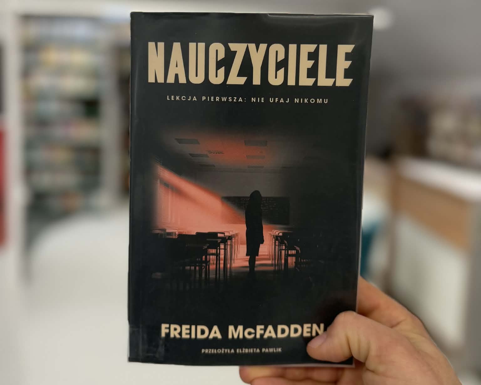 Na pierwszym planie widnieje męska dłoń trzymająca książkę Freidy McFadden "Nauczyciele". W tle widać rozmyte regały z książkami. Zdjęcie zrobione jest w bibliotece
