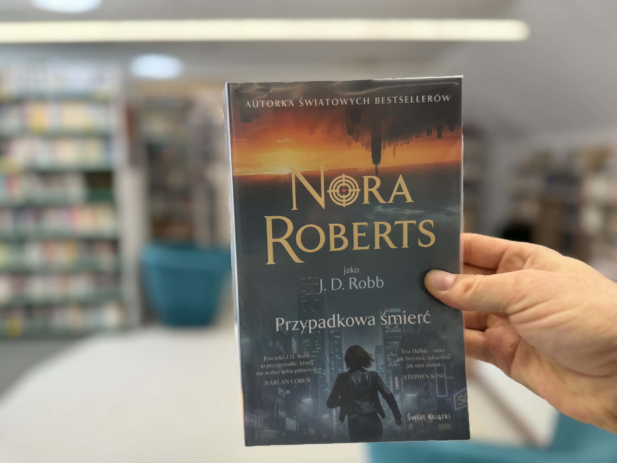Na pierwszym planie widnieje męska dłoń trzymająca książkę Nory Roberts "Przypadkowa śmierć". W tle widać rozmyte regały z książkami. Zdjęcie zrobione jest w bibliotece