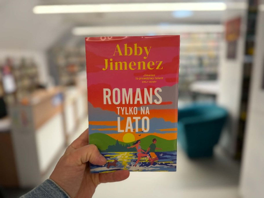 Na pierwszym planie osoba trzyma w dłoni książkę „Romans tylko na lato” autorstwa Abby Jimenez. Okładka jest utrzymana w intensywnych kolorach czerwieni, pomarańczu i błękitu, z ilustracją dwojga ludzi nad jeziorem o zachodzie słońca. W tle, lekko rozmytym, widoczne jest wnętrze biblioteki z regałami na książki