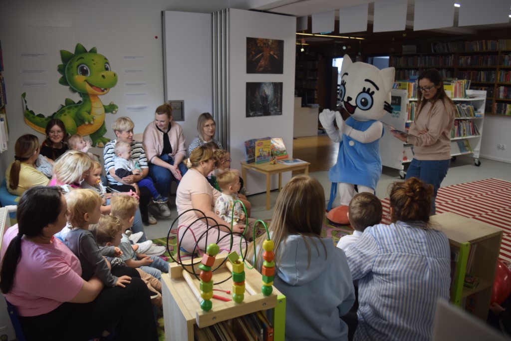 Grupa małych dzieci wraz z opiekunami siedzi w kręgu w kąciku dziecięcym w bibliotece. Wszyscy z uwagą patrzą na postać w dużym kostiumie maskotki Kici Koci, która stoi przed nimi w niebieskiej sukience i białych rękawiczkach. Obok maskotki stoi kobieta trzymająca otwartą książkę. Na ścianie po lewej stronie widnieje duża naklejka z zielonym smokiem, a na podłodze leży kolorowa mata z zabawkami