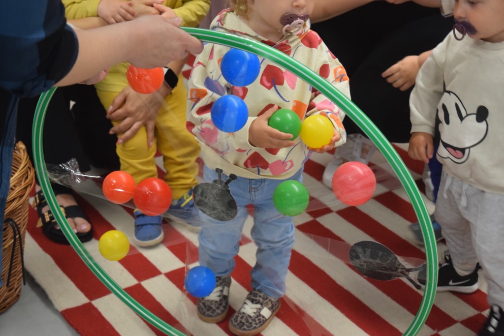 Maluchy uczestniczące w zabawie ruchowej. Dziecko w jasnej bluzie trzyma plastikowe piłeczki przed zielonym obręczem hula-hoop, na który naklejone są kolorowe elementy i ilustracje owadów