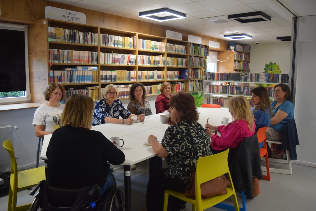 Grupa ośmiu kobiet siedzi przy dużym, białym stole w nowoczesnej sali bibliotecznej. Kobiety rozmawiają, piją kawę i herbatę. W tle widoczne są wysokie, drewniane regały wypełnione książkami oraz kolorowe krzesła. Jedna z uczestniczek porusza się na wózku 