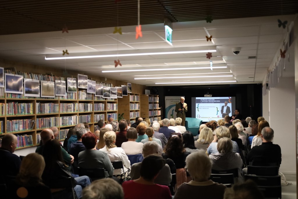 Szerokie ujęcie sali bibliotecznej wypełnionej publicznością podczas spotkania autorskiego. Na końcu sali, na podwyższeniu, stoi Jacek Fedorowicz z mikrofonem w ręku. W tle znajduje się ekran z napisem „Spotkanie autorskie z Jackiem Fedorowiczem”. Wzdłuż lewej ściany ciągną się wysokie regały z książkami
