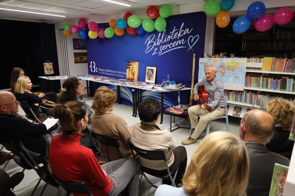 Widok z góry na kameralne spotkanie autorskie w bibliotece. Podróżnik Andrzej Ciuba siedzi przy regale z książkami i gra na gitarze klasycznej, a przed nim siedzi na krzesłach wpatrzona publiczność. Na stołach w tle wyłożone są pamiątki i zdjęcia z wypraw