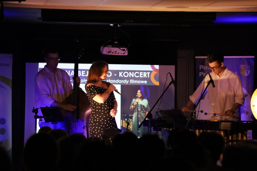 Koncert zespołu Joanna Bejm Trio widziany z perspektywy publiczności, której zaciemnione sylwetki znajdują się na pierwszym planie. Na oświetlonej scenie występuje wokalistka w ciemnej sukience w grochy, śpiewając do mikrofonu. Po jej lewej stronie mężczyzna gra na kontrabasie, a po prawej drugi muzyk gra na wibrafonie. W tle wyświetlany jest napis zapowiadający najpiękniejsze standardy filmowe