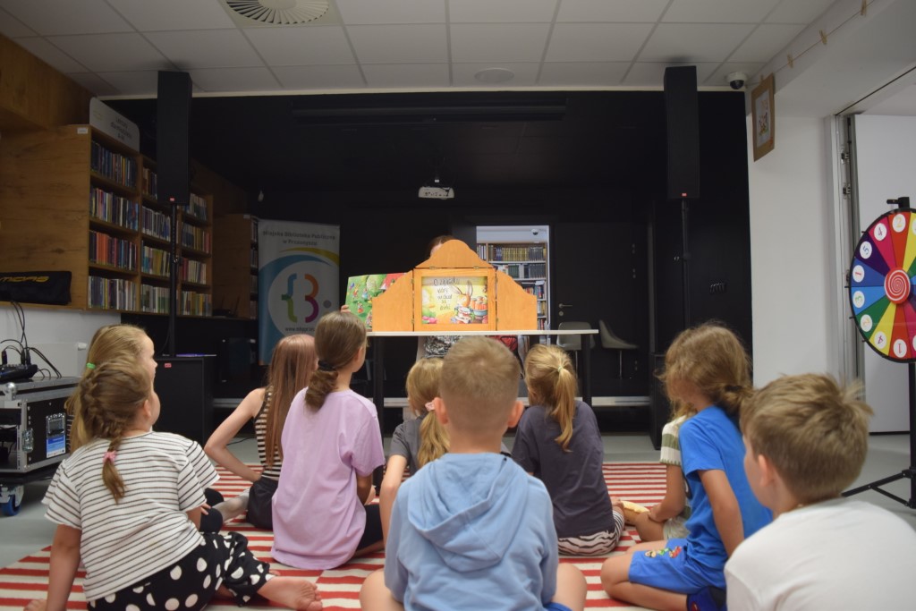 Dzieci siedzą tyłem na pasiastym dywanie, oglądając przedstawienie prezentowane w drewnianej skrzynce teatrzyku Kamishibai, która stoi na stole. W tle widać regał z książkami i kolorowe koło fortuny