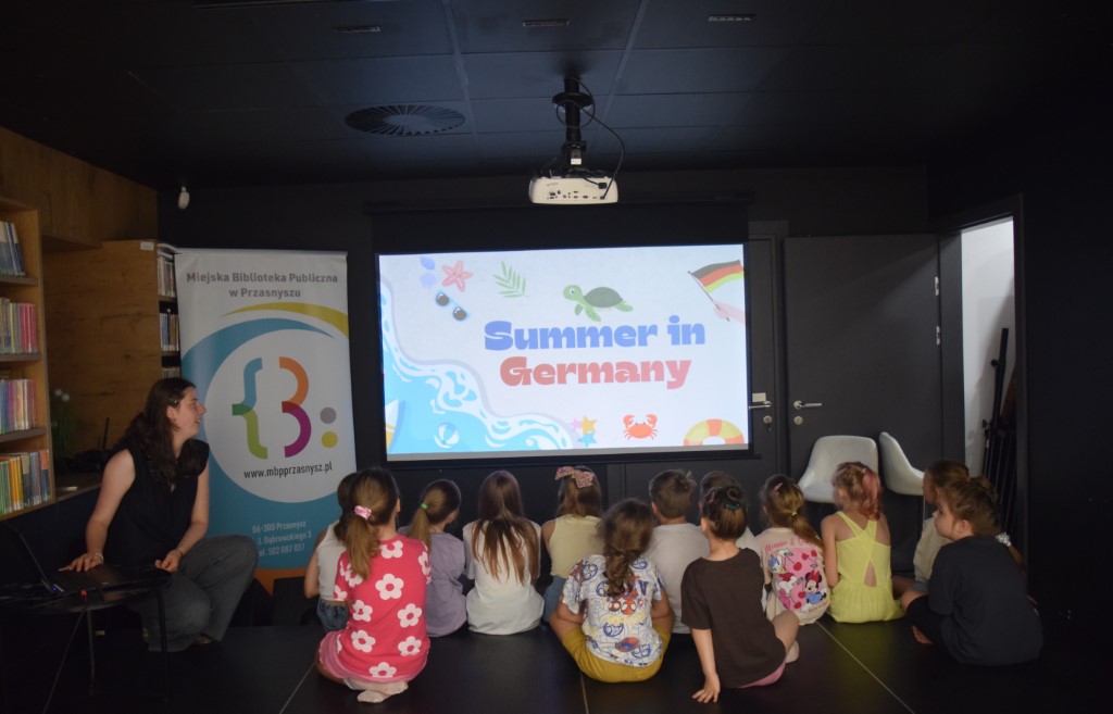 Grupa dzieci siedzi tyłem na podłodze, oglądając wyświetlany na dużym ekranie slajd z napisem „Summer in Germany” i rysunkiem żółwia morskiego. Po lewej stronie na podwyższeniu siedzi prowadząca zajęcia, obok której stoi pionowy baner reklamowy biblioteki