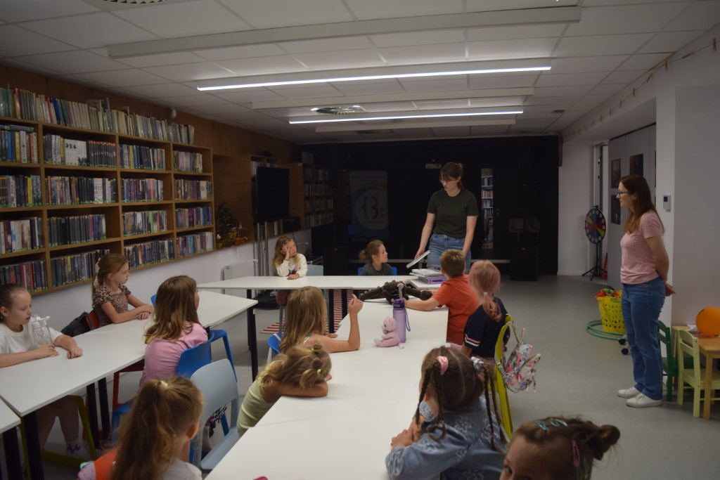 Grupa dzieci siedzi przy białych stołach ustawionych w salce bibliotecznej, słuchając stojącej przed nimi prowadzącej w ciemnozielonej koszulce. Z boku przygląda się im druga opiekunka, a po lewej stronie widać regały pełne książek
