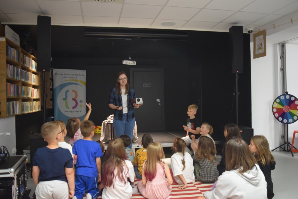 Kobieta w rozpiętej koszuli w kratę stoi na niskim podwyższeniu w sali bibliotecznej i trzyma retro projektor, przemawiając do grupy dzieci siedzących przed nią tyłem na pasiastym dywanie