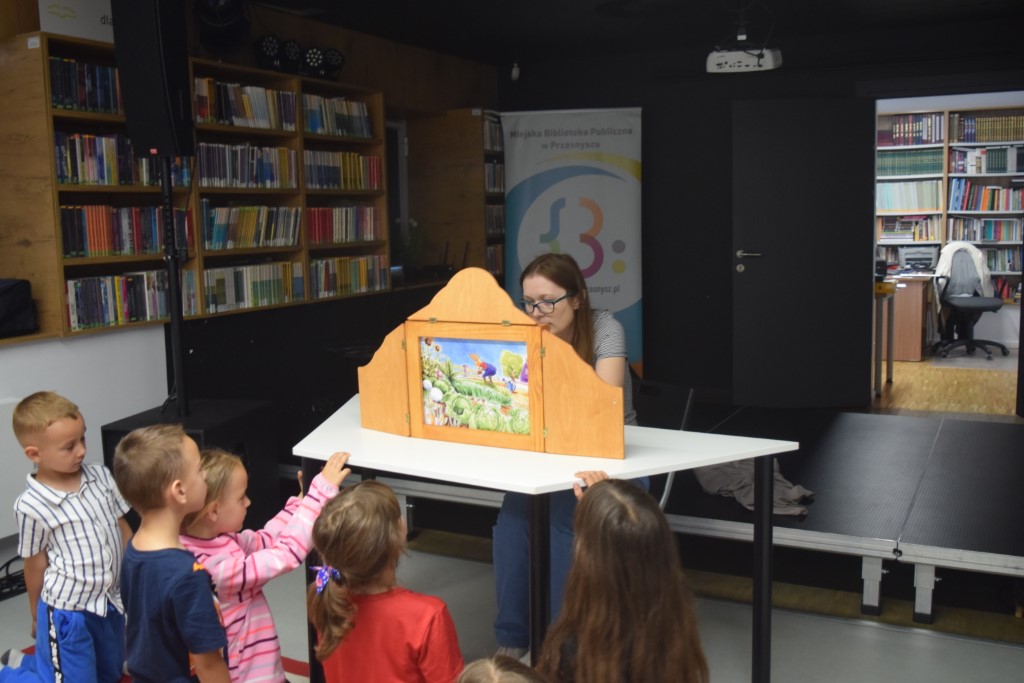 Kobieta w okularach prezentuje ilustrację w drewnianej skrzynce teatrzyku Kamishibai, przed którą stoi i przygląda się grupa małych dzieci. W tle widać regały z książkami oraz wejście do sąsiedniego pomieszczenia biblioteki