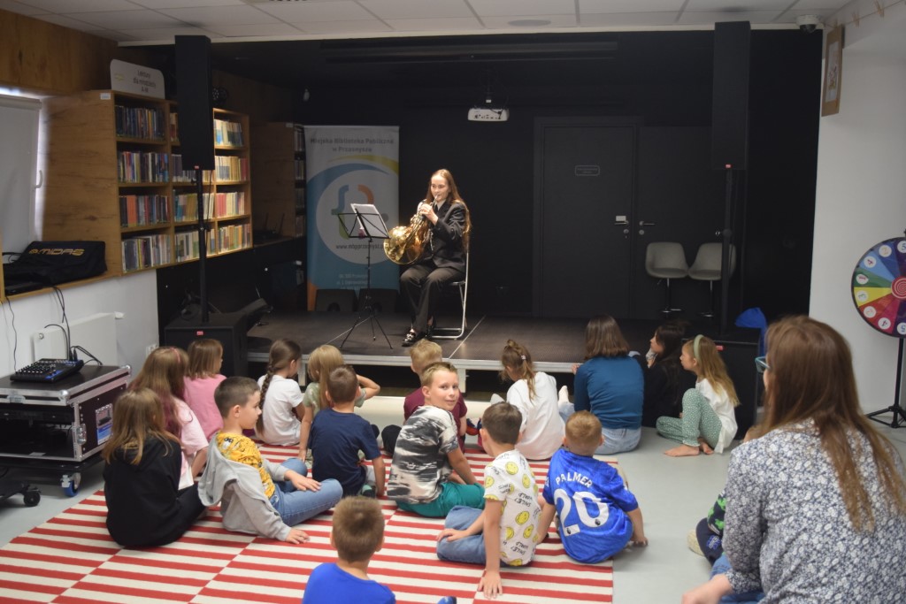 Dziewczyna w ciemnym garniturze gra na waltorni, siedząc przy pulpicie na małej scenie w bibliotece. Przed nią, na pasiastym dywanie, siedzi grupa dzieci słuchających koncertu