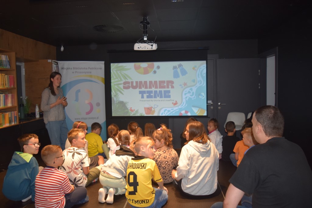 Prowadząca zajęcia stoi przy ekranie projekcyjnym wyświetlającym slajd z napisem „SUMMER TIME”. Na podłodze siedzi grupa dzieci – jedno z nich ma na sobie żółtą koszulkę piłkarską z numerem 9 i nazwiskiem Lewandowski