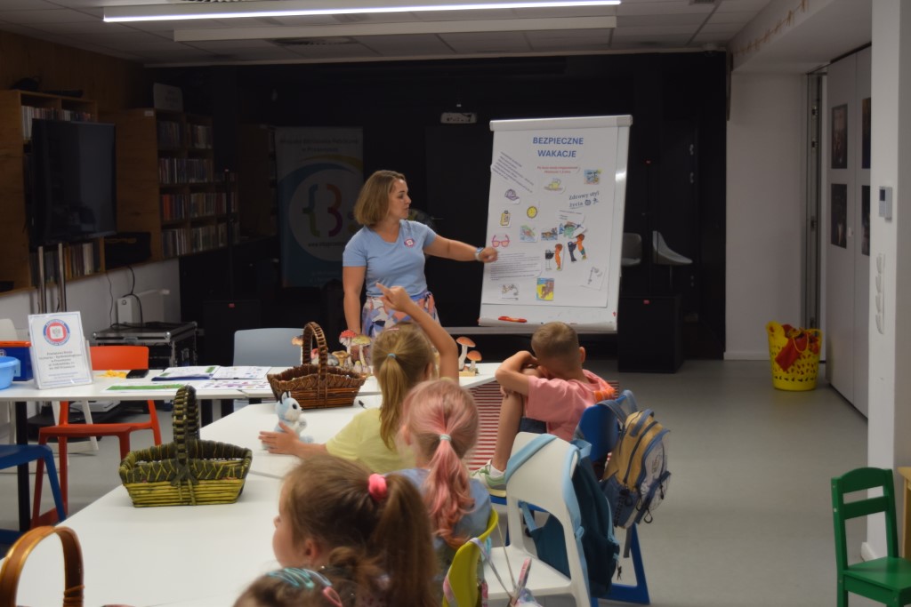 Kobieta stoi przy tablicy flipchart z napisem „Bezpieczne wakacje” i wskazuje na przypięte do niej rysunki edukacyjne. Przed nią, przy białym stole, siedzi tyłem grupa dzieci