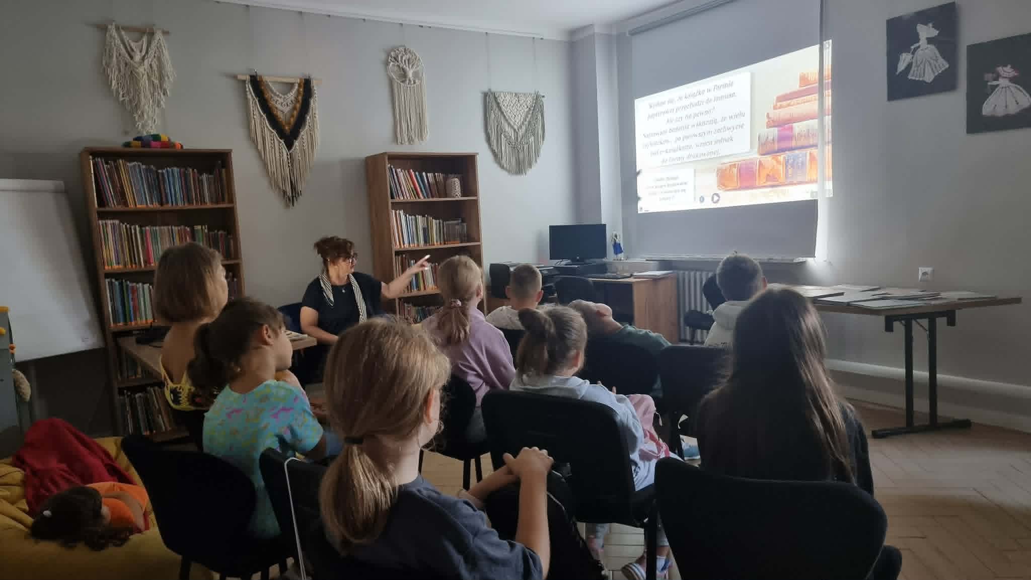 Grupa dzieci siedzi na krzesłach w sali bibliotecznej, oglądając prezentację wyświetlaną z projektora na ścianie. Prowadząca w okularach stoi przy regale z książkami i wskazuje dłonią na ekran