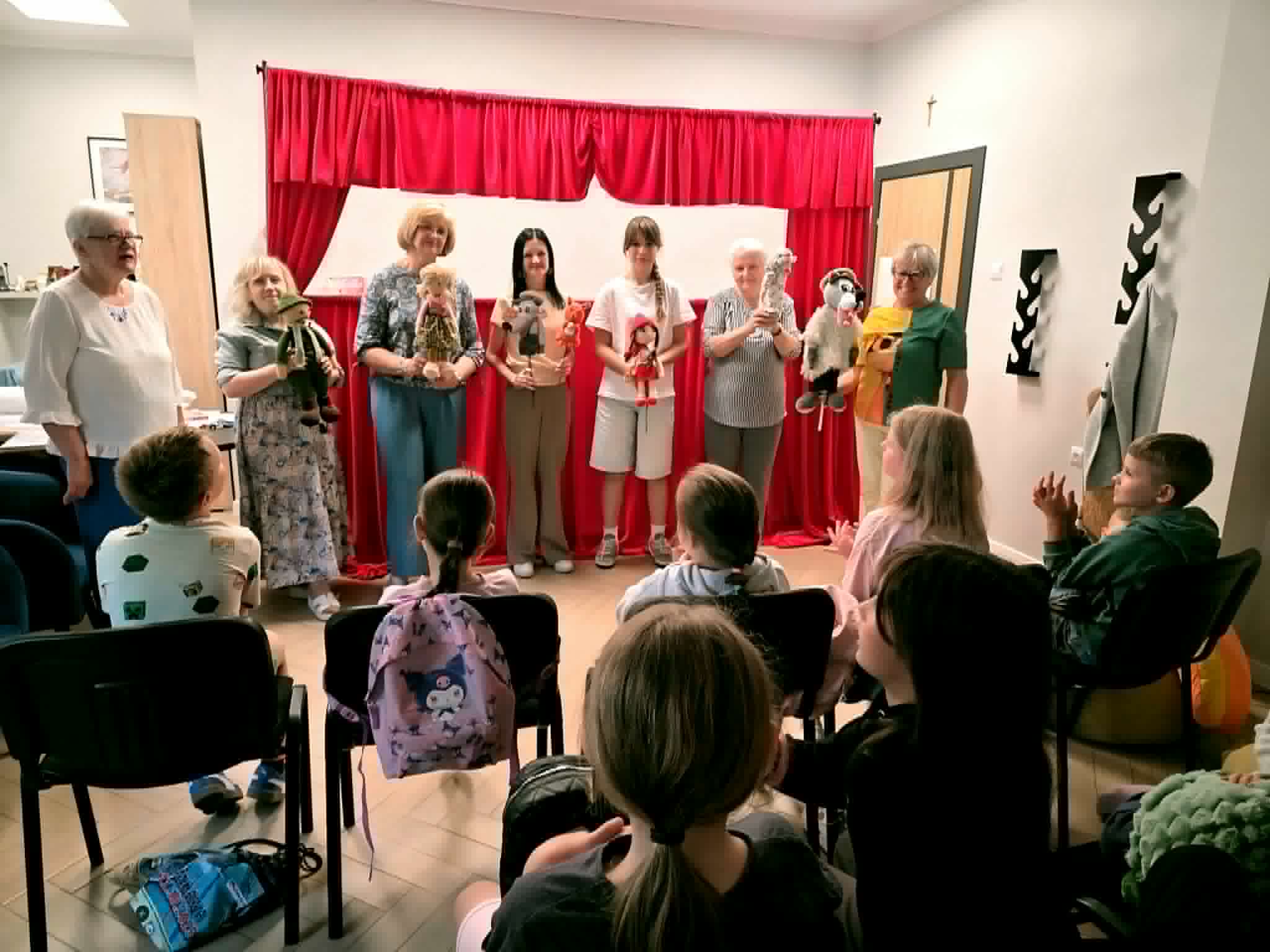 Grupa seniorek stoi na scenie za czerwoną kurtyną teatrzyku kukiełkowego, prezentując dzieciom trzymane w rękach pacynki przedstawiające postaci z bajki o Czerwonym Kapturku. Widownia siedząca tyłem na krzesłach obserwuje występ