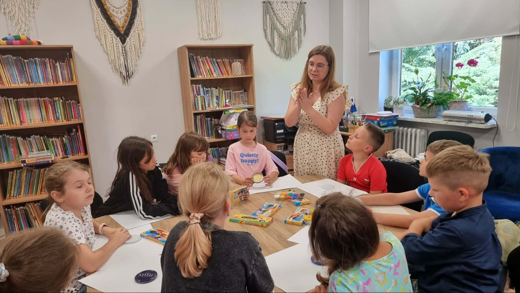 Grupa dzieci siedzi przy drewnianym stole w jasnej sali bibliotecznej i słucha stojącej przed nimi instruktorki w okularach oraz jasnej sukience w groszki, która gestykuluje dłońmi. Na stole leżą opakowania z plasteliną i białe kartki papieru