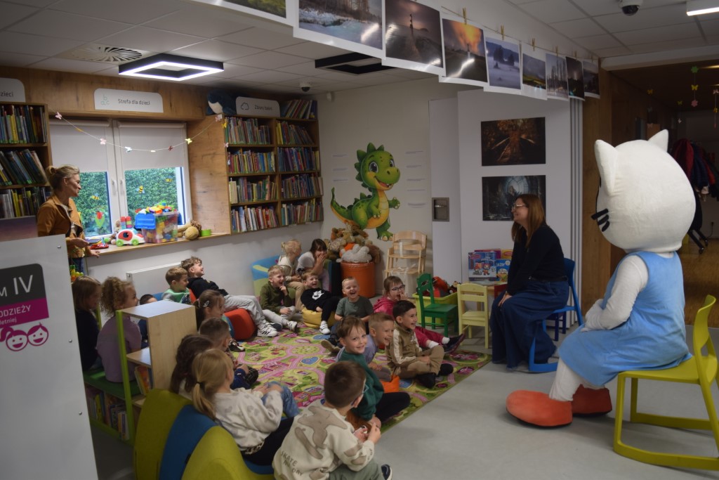 Bibliotekarka w okularach siedzi na niskim krześle i rozmawia z grupą dzieci siedzących na kolorowym dywanie w strefie dla najmłodszych. Obok niej na żółtym krześle siedzi duża maskotka Kici Koci. Dzieci słuchają z zaciekawieniem, a w tle widać regały z zabawkami i książkami oraz wystawę zdjęć