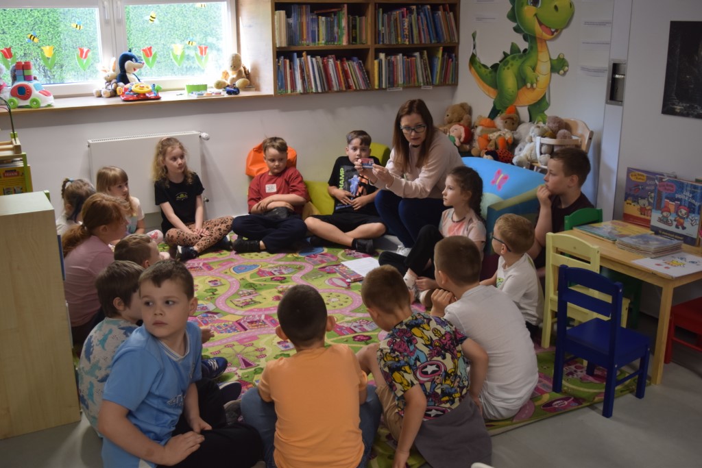 Dzieci siedzą w kręgu na wzorzystym, kolorowym dywanie w kąciku dziecięcym w bibliotece. Przed nimi kuca kobieta w okularach, która trzyma w dłoniach małą kartkę i pokazuje ją grupie. W tle widać niskie regały z książkami, pluszowe zabawki na parapecie okna oraz dużą naklejkę z zielonym smokiem na ścianie