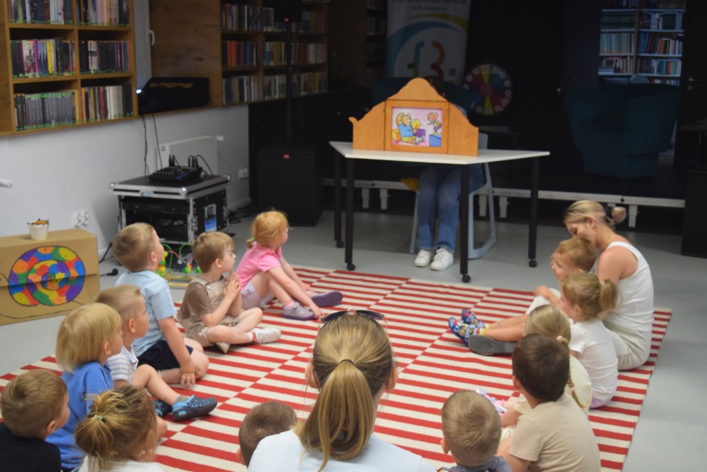 Grupa małych dzieci wraz z opiekunami siedzi na pasiastym, czerwono-białym dywanie w sali bibliotecznej. Wszyscy z zaciekawieniem oglądają teatrzyk obrazkowy Kamishibai, prezentowany w drewnianej skrzynce na stoliku przed nimi. W tle po lewej stronie stoją regały z książkami