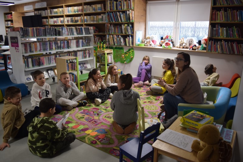 Zdjęcie przedstawia grupę dzieci, które siedzą przy białych stolikach. wszystkie są zwrócone do bibliotekarek, które prowadzą zajęcia