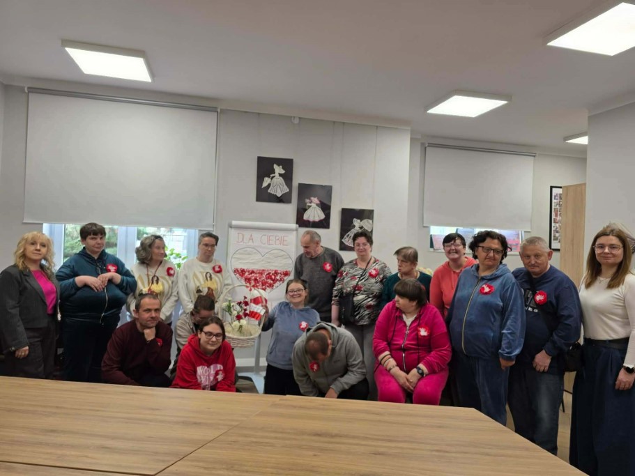 Grupa kilkunastu uśmiechniętych osób pozuje do wspólnego zdjęcia w jasnej sali. Niektórzy mają przypięte do ubrań czerwono-białe kotyliony. W tle stoi tablica prezentacyjna z napisem „DLA CIEBIE” i narysowanym sercem wypełnionym biało-czerwonymi elementami. Przed grupą stoi duży, drewniany stół