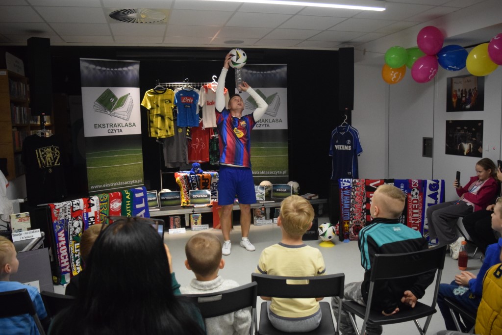 Mężczyzna w koszulce FC Barcelony wykonuje efektowną sztuczkę, balansuąc piłką nożną umieszczoną na plastikowej butelce nad głową. Zgromadzona na krzesłach widownia przygląda się pokazowi